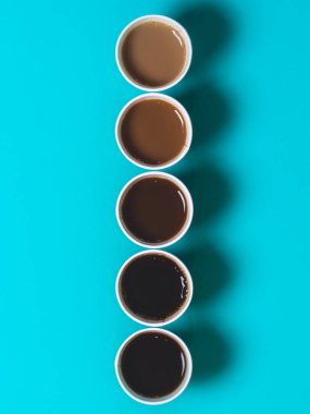 Kahveye farklı miktarlarda süt eklenerek renk gradyanı oluşturacak şekilde dizilmiş minimalist kahve fincanları. Turkuaz arkaplan, düz üst görünüm
