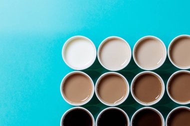 Kahveye farklı miktarlarda süt eklenerek renk gradyanı oluşturacak şekilde dizilmiş minimalist kahve fincanları. Turkuaz arkaplan, düz üst görünüm