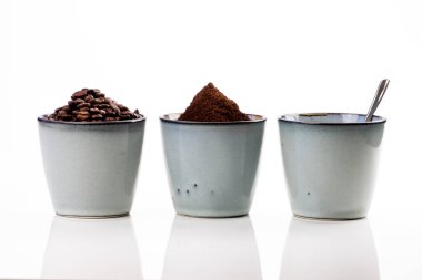 Sıcak kahve, fasulye ve öğütülmüş kahveyle dolu 3 tane köy kahvesi. Hafif yansıtıcı bir masanın üzerinde duran fincanlar beyaz bir arkaplanda kaybolur.