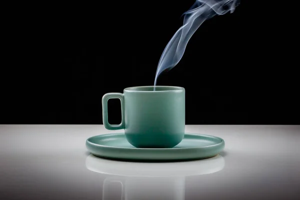 Pastel green colored coffee or tea cup with hot liquid, smoke and steam, black background. Cup standing on a slightly reflective table that fades into a black background