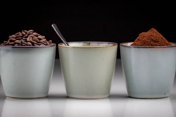 3 rustic coffee cups with coffee grounds,table coffee, and ground coffee. Standing on a white reflective table fading into a black background