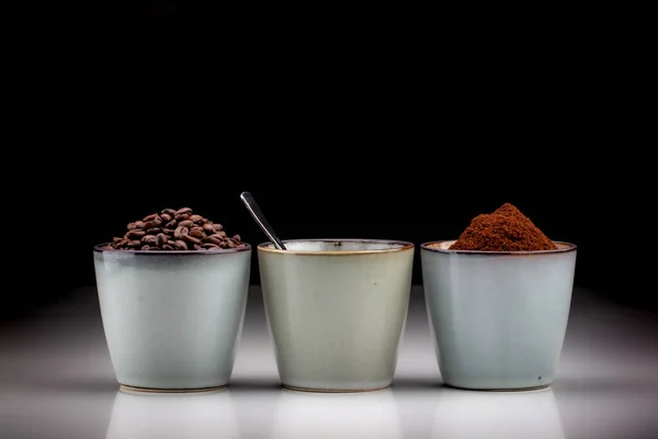 3 rustic coffee cups with coffee grounds,table coffee, and ground coffee. Standing on a white reflective table fading into a black background