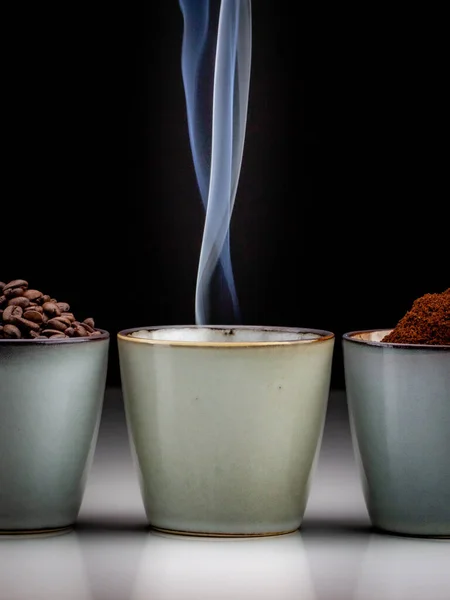 3 rustic coffee cups with coffee grounds, smoking coffee, and ground coffee. Standing on a white reflective table fading into a black background