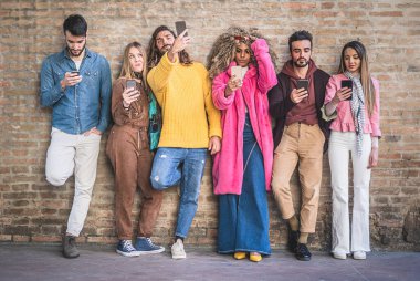 Bir grup çok ırklı genç, akıllı telefon ve sosyal medya müptelası tuğla duvarın üzerinde duruyor. Milenyum insanları akıllı telefonlar ve yeni teknolojiler kullanıyor.