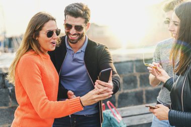 Bir grup insan güneş gözlüğü takıyor, akıllı telefon ekranını izliyor ve birlikte gülüyorlar. Gençler yeni teknoloji kavramını kullanıyor.