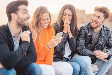 Dört arkadaş grubu birlikte eğleniyor. İki kadın ve iki erkek bankta oturmuş gülüyor ve akıllı telefon kullanarak eğleniyorlar..