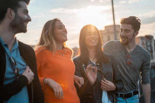 Group of trendy young people chatting together standing on a wall outdoors. Students having fun together. Focus on on the blonde girl smiling. Lifestyle concept.