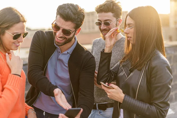 Group of young people laughing watching a joke or a video on a smartphone screen. All of them wearing sunglasses. Orange sunset tones, lifestyle technology concept.
