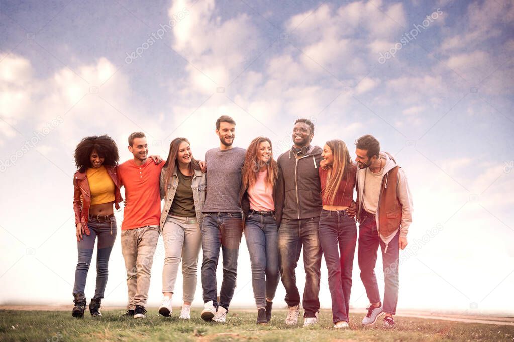 Group of diverse young people walking arm around shoulders. Millennial generation teamwork having fun bonding talking together against a cloudy sky. Collaboration, help and carefree lifestyle concept.