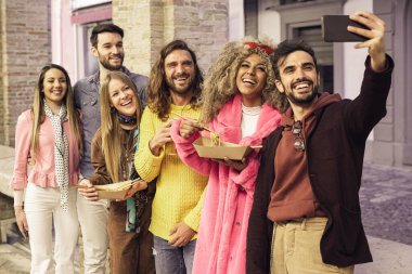 Genç çoklu etnik gruplar akıllı telefonlarıyla erişte yerken selfie çekiyorlar. Gençler dışarıda Çin yemeği yiyor ve mobil cihazlarla eğleniyorlar..