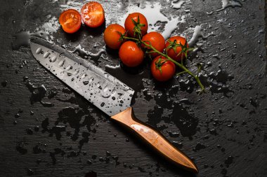 Santoku bıçağı ıslak siyah bir kesme tahtasıyla kiraz domatesleri kesiyor. Yukarıdan görüntüle.
