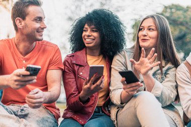 Bir grup modaya uygun genç ellerinde akıllı telefonlar, dışarıda bir bankta oturup sohbet ediyorlar. Öğrenciler birlikte eğleniyor. Ağzını açıp gülümseyen Afro-Amerikalı bir kıza odaklan.