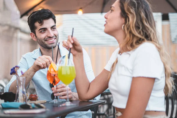 Smiling couple of happy young people drinking cocktails sitting on an outdoors restaurant in the summer.  Millennials relaxing drinking alcoholic drinks concept.