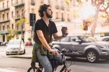 Yetişkin sakallı hippi, şehirde bisiklet sürerken kahve içiyor. Sağlık hayatı ve yaşam tarzı kavramı.