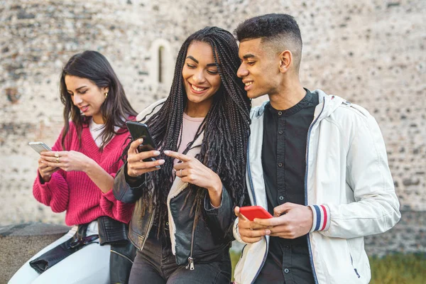 Gen Z young people using smartphone and social networks together. Multiracial people having fun together watching cell phone screens outdoors.