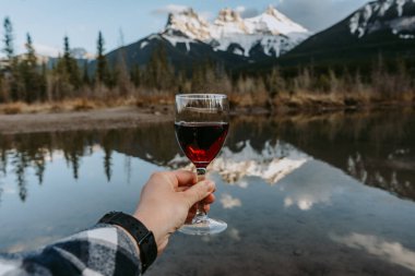 Canmore, Alberta 'da Polis Deresi' nde bir kadeh kırmızı şarap tutan bir kadın eli. Üç Kız Kardeş Dağı 'nın zirveleri arka plandaki suya yansıyor. Kanada Kayalıkları.