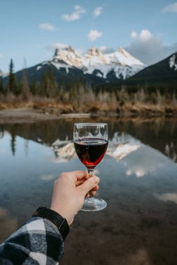 Canmore, Alberta 'da Polis Deresi' nde bir kadeh kırmızı şarap tutan bir kadın eli. Üç Kız Kardeş Dağı 'nın zirveleri arka plandaki suya yansıyor. Kanada Kayalıkları.