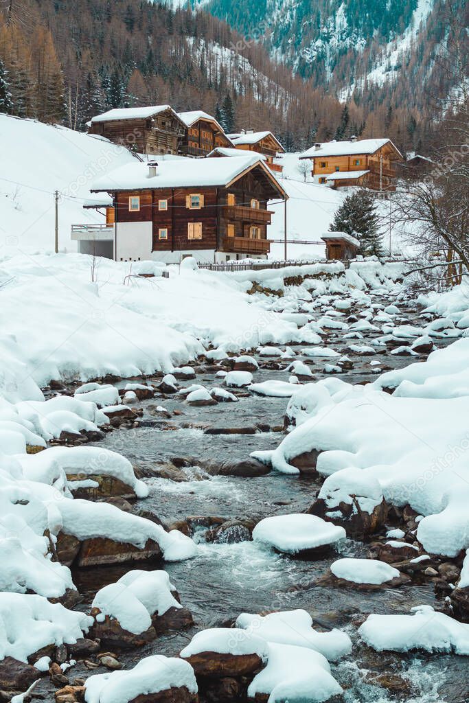 Ice and snow covered creek on a cold winter day infront of traditional wooden cabins of little mountain village St. Gertraud in Ultental, South Tyrol/Italy