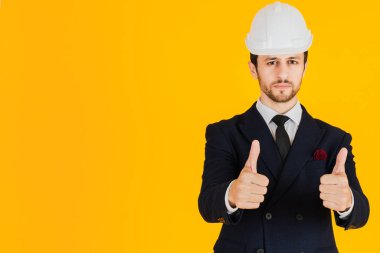 Takım elbiseli genç bir mühendis ve beyaz bir güvenlik bölmesi giyiyor. Takım elbiseli genç bir mimarın stüdyosunda portresi ve sarı izole edilmiş bir kask giyiyor. Pankartlar için resim.