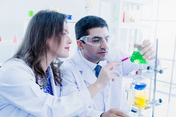 一位中东的男性科学家和一位高加索的美丽女性科学家正在一个科学实验室里分析闪光中的绿色溶液 — 图库照片