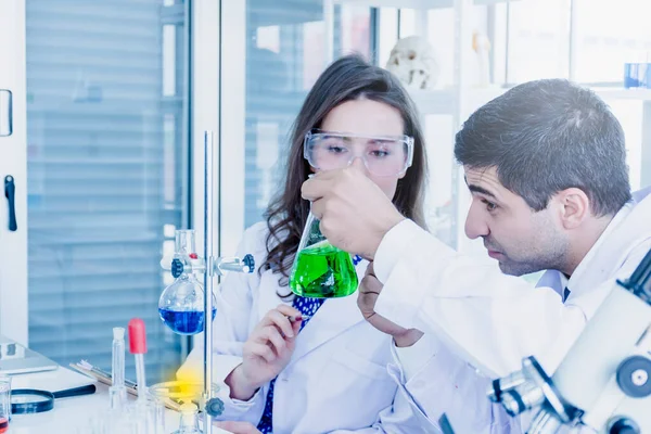一位中东的男性科学家和一位高加索的美丽女性科学家正在一个科学实验室里分析闪光中的绿色溶液 — 图库照片