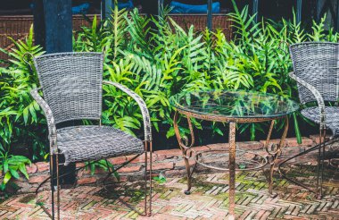 Eski bir metal masa ve kahve dükkanının dışında arka planda eğreltiotu ve cam bina duvarı bulunan iki sandalye bulunuyor. Metal gövdeli sandalyeler ve hasır örgüler.