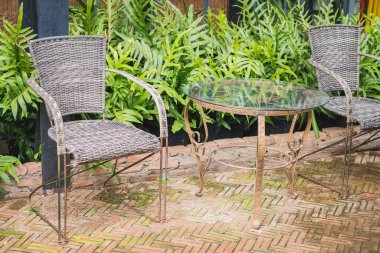 Eski bir metal masa ve kahve dükkanının dışında arka planda eğreltiotu ve cam bina duvarı bulunan iki sandalye bulunuyor. Metal gövdeli sandalyeler ve hasır örgüler.