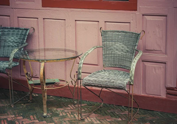 A vintage metal table and 2 chairs are on the outside of the coffee shop, with a wooden wall of building in the background vintage. Chairs with metal frame and wicker weave as the body.