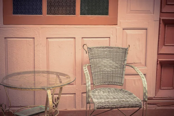 A vintage metal table and chair are on the outside of the coffee shop, with a wooden wall of building in the background vintage. Chair with metal frame and wicker weave as the body.