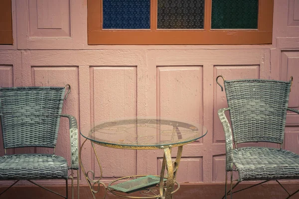 A vintage metal table and 2 chairs are on the outside of the coffee shop, with a wooden wall of building in the background vintage. Chairs with metal frame and wicker weave as the body.