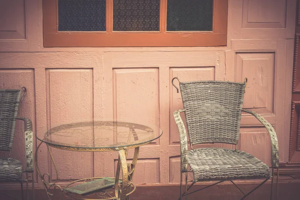 A vintage metal table and 2 chairs are on the outside of the coffee shop, with a wooden wall of building in the background vintage. Chairs with metal frame and wicker weave as the body.