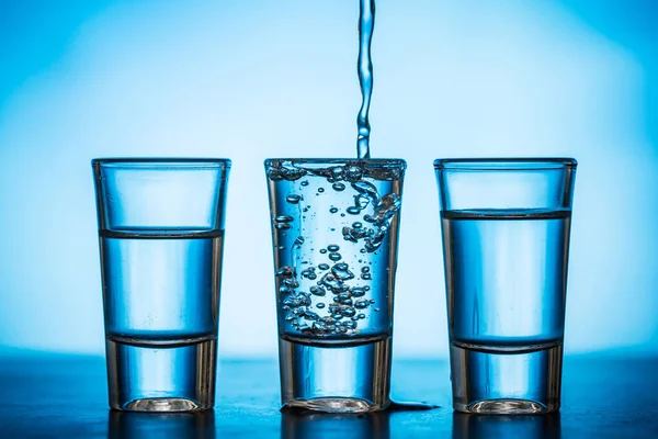 Água Sendo Derramada Pequenos Copos Fundo Azul — Fotografia de Stock
