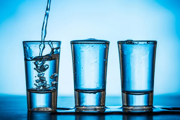 Água Sendo Derramada Pequenos Copos Fundo Azul — Fotografia de Stock
