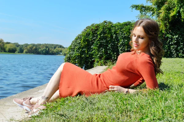 Jong meisje in de oranje jurk zit op het gras bij het water. — Stockfoto