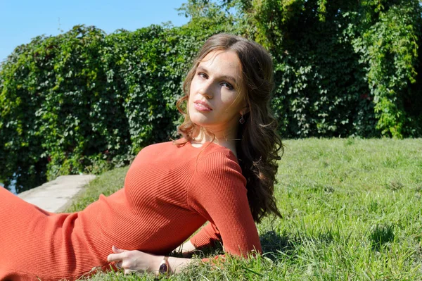Turuncu elbiseli genç kız suyun yanındaki çimlerin üzerinde oturuyor.. — Stok fotoğraf