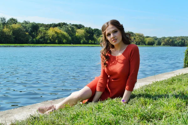 Narancssárga ruhás fiatal lány ül a fűben a víz mellett.. — Stock Fotó