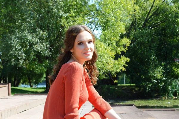 Chica joven en vestido naranja se sienta en un muelle cerca del agua . — Foto de Stock