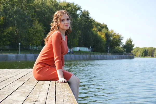 Молодая девушка в оранжевом платье сидит на пирсе у воды . — стоковое фото