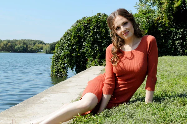 Young girl in the orange dress is sitting on the grass near the water. — 스톡 사진