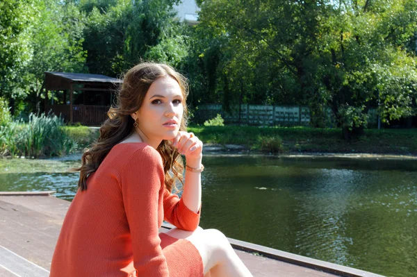 Młoda dziewczyna w pomarańczowej sukience siedzi na molo w pobliżu wody. — Zdjęcie stockowe