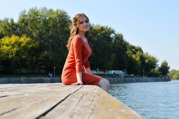 Turuncu elbiseli genç kız su kenarında bir iskelede oturuyor.. — Stok fotoğraf