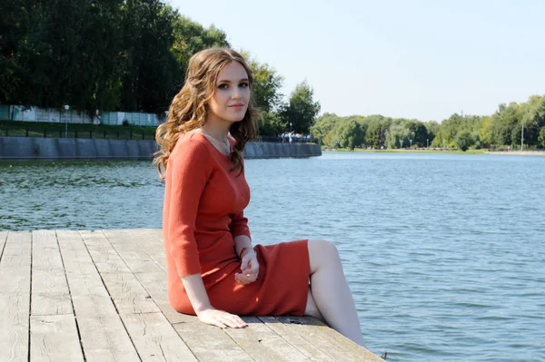 Chica joven en vestido naranja se sienta en un muelle cerca del agua . —  Fotos de Stock