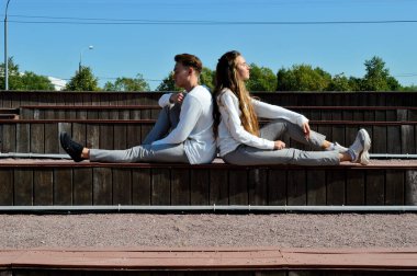 Genç bir kız ve aynı kıyafetleri giyen genç bir adam ahşap bir bankta oturuyor..