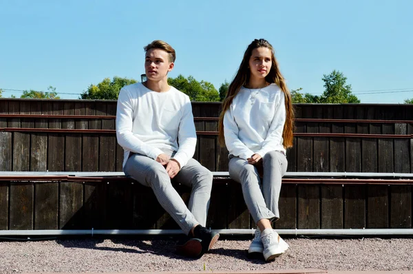Młoda dziewczyna i młody mężczyzna w tych samych ubraniach siedzący na drewnianej ławce. — Zdjęcie stockowe