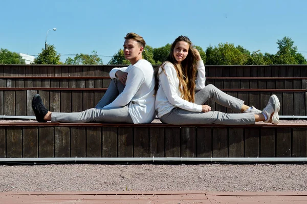 Mladá dívka a mladý muž ve stejném oblečení sedí na dřevěné lavičce. — Stock fotografie