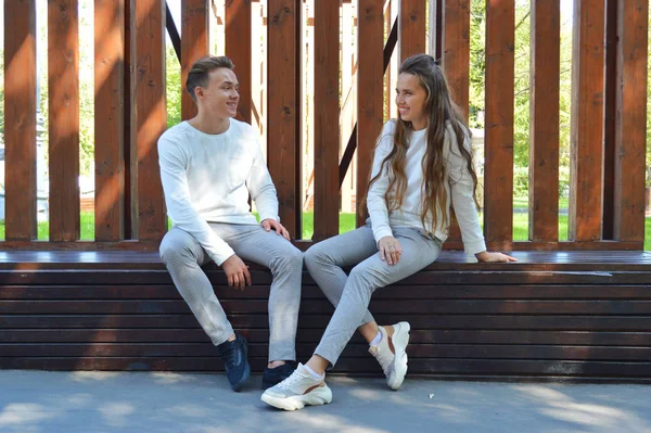 En ung flicka och en ung man i samma kläder sitter på en träbänk. — Stockfoto