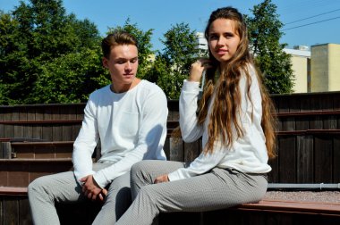 Genç bir kız ve aynı kıyafetleri giyen genç bir adam ahşap bir bankta oturuyor..