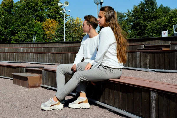 Uma jovem e um jovem com as mesmas roupas sentados em um banco de madeira . — Fotografia de Stock