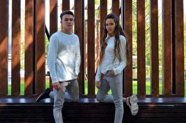 Genç bir kız ve aynı kıyafetleri giyen genç bir adam ahşap bir bankta oturuyor..