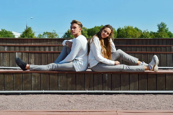 Uma jovem e um jovem com as mesmas roupas sentados em um banco de madeira . — Fotografia de Stock